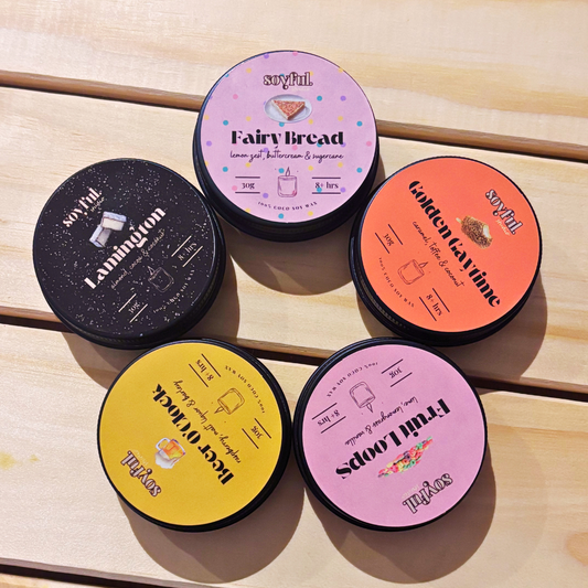 Five small candle tins with colorful lids featuring text and graphics on a wooden surface.